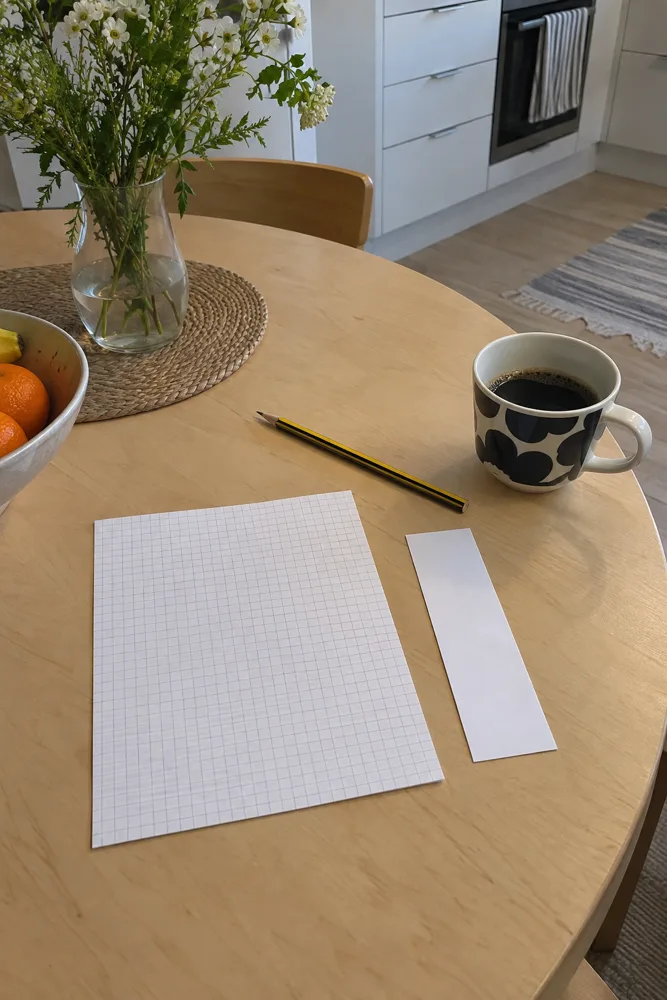 Tyhjä ruutupaperi, kapea paperilappu, lyijykynä ja kahvikuppi koivuvanerisella pöydällä